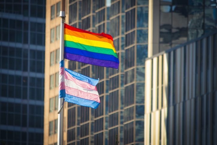 Pride and Trans Flag waving in the wind in a downtown urban setting. Office buildings can be seen behind.